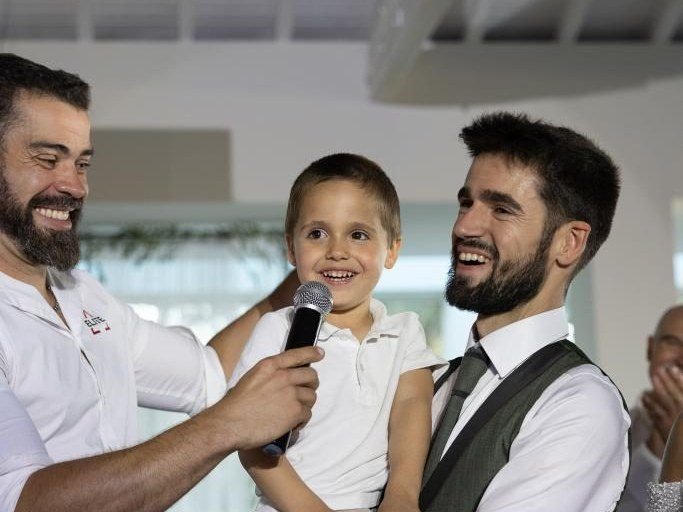 Criança ao colo, com um microfone na mão, a sorrir com dois adultos numa festa de batizado
