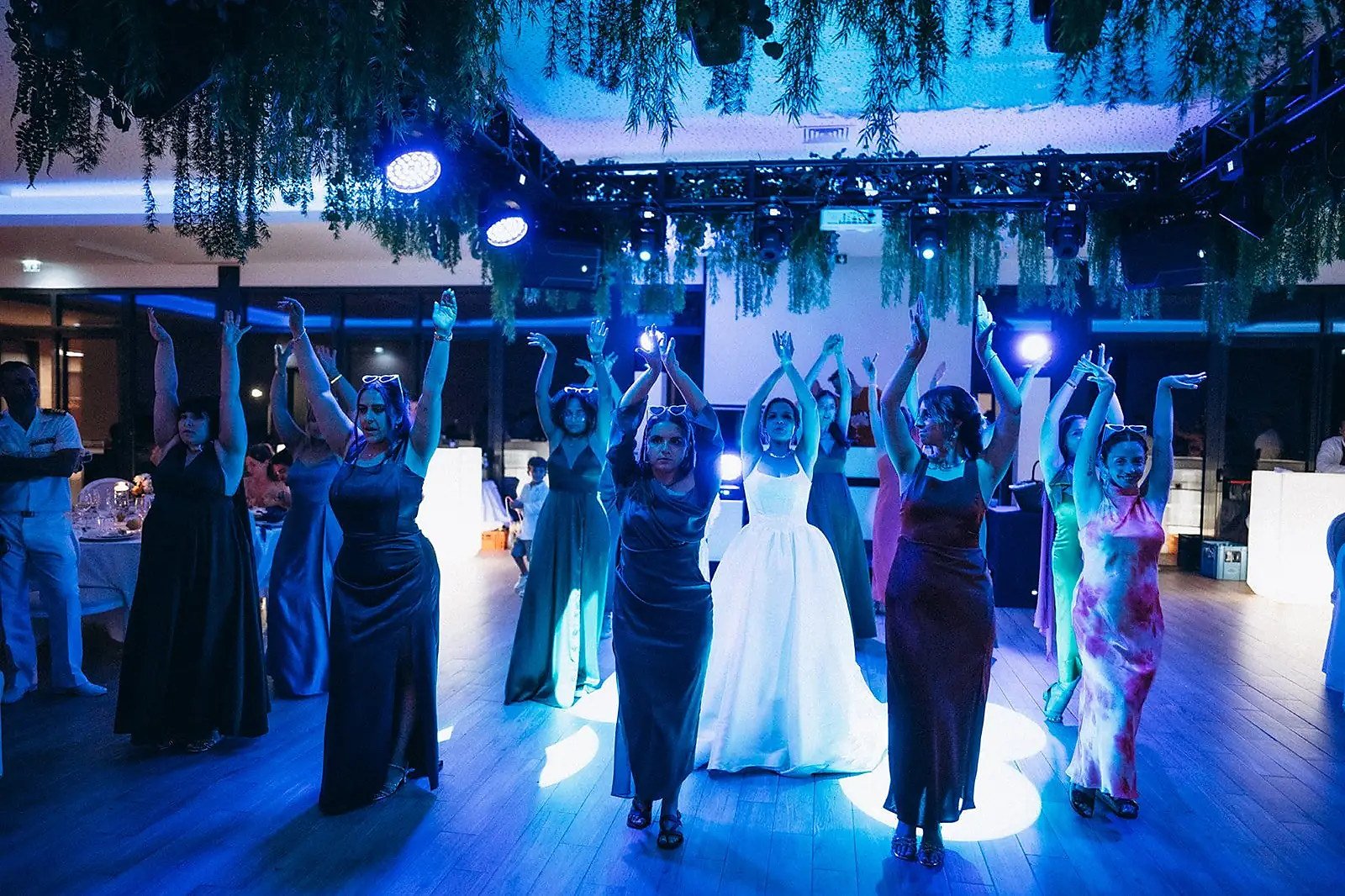Convidados de braços no ar numa coreografia de grupo, pista iluminada a azul