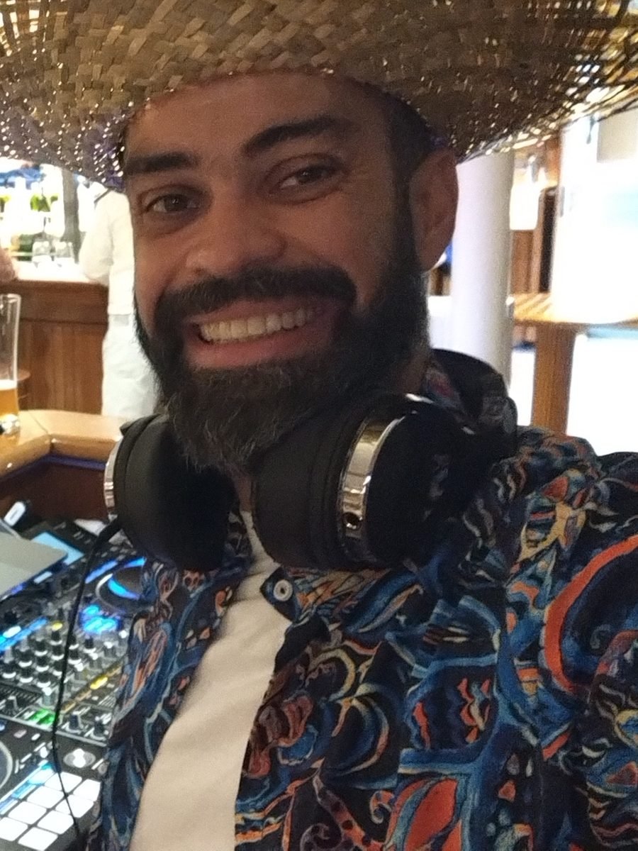 Guga Brasa de chapéu de palha e camisa florida, a sorrir na cabine de DJ, com os auscultadores ao pescoço